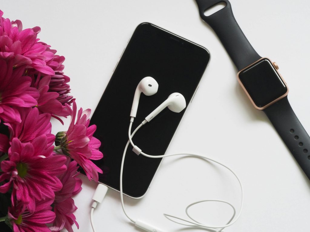 A flat lay of smartphone, smartwatch, and earphones beside vibrant pink flowers on a white background.